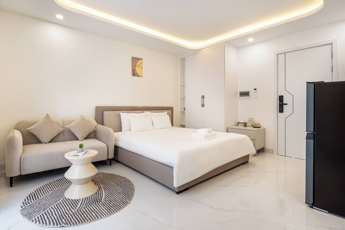 A modern hotel room with a bed, a sofa, a small table, and a refrigerator.