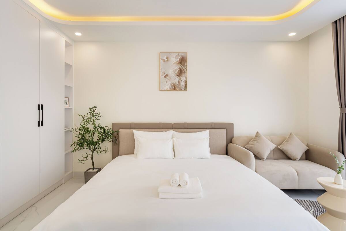 A modern hotel room with a large bed, a sofa, a small table, and a potted plant.
