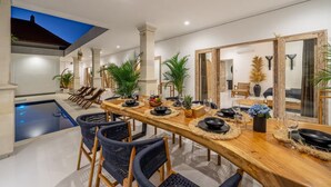 Uma sala de jantar com uma mesa de madeira comprida, cadeiras e uma piscina ao fundo.