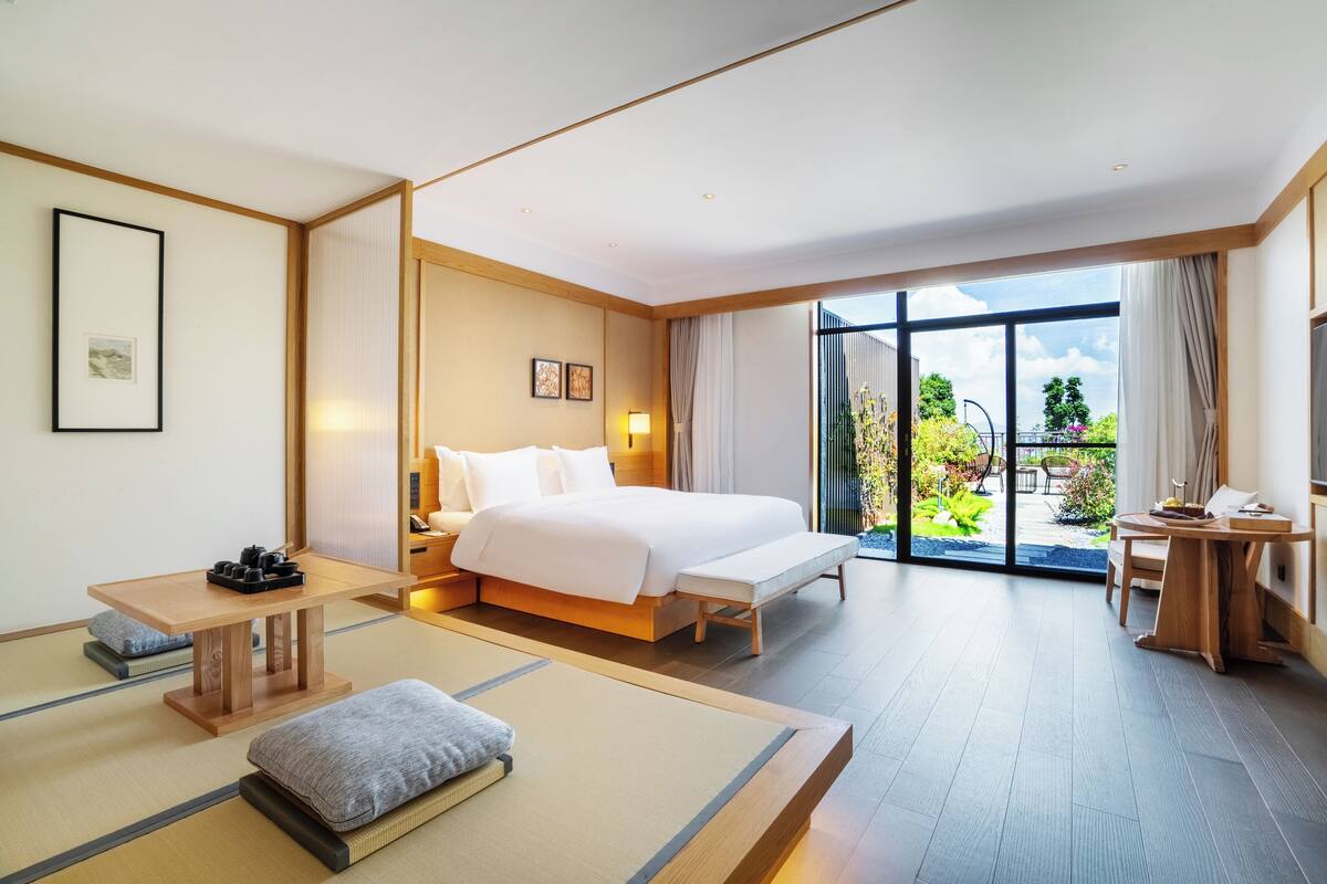 Habitación de hotel con cama, escritorio, banco y vista a un jardín a través de amplios ventanales.