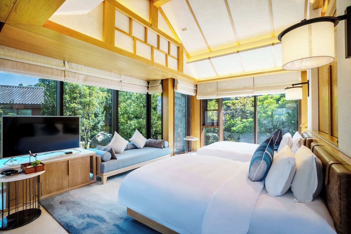 Habitación de hotel moderna con una cama grande, un televisor, una zona de estar con cojines y ventanas amplias con vistas a la vegetación.