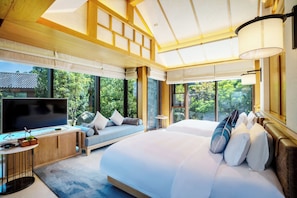 Ein modernes Hotelzimmer mit einem großen Bett, einem Fernseher, einer Sitzecke mit Kissen und großen Fenstern mit Blick auf Grünflächen.