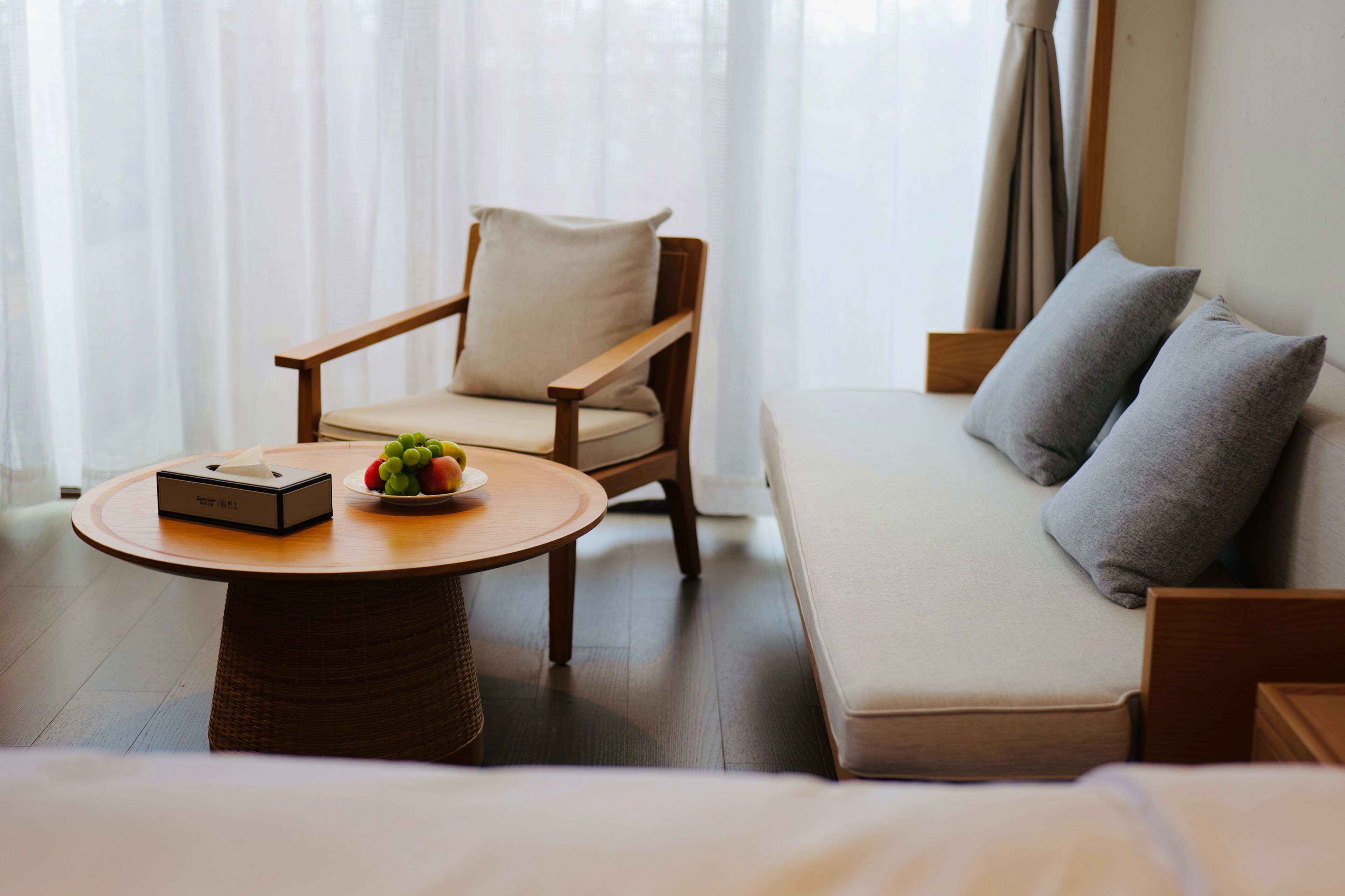 Una habitación con una cama, una silla de madera, una mesa redonda con una caja y fruta, y un sofá con almohadones.
