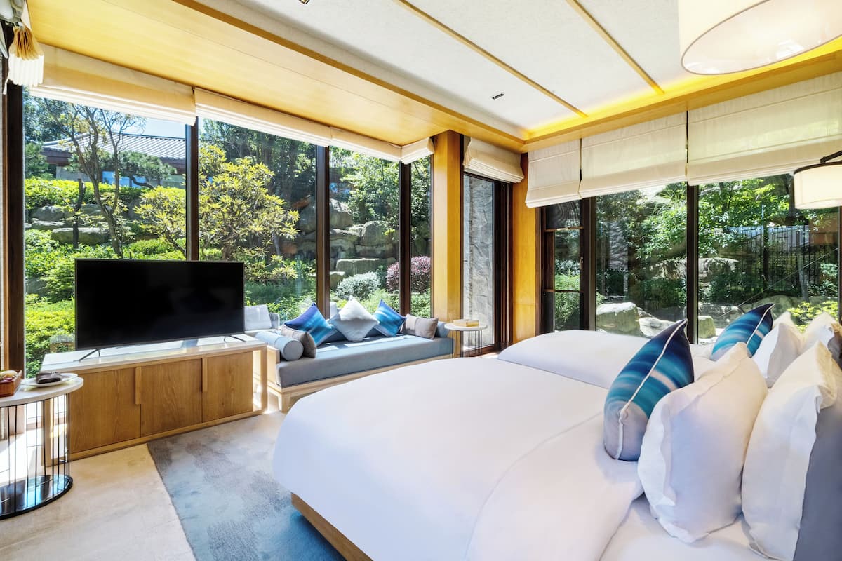 Habitación de hotel moderna con una cama grande, televisión, zona de estar con vistas al jardín y ventanas amplias.