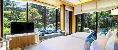 Habitación de hotel moderna con una cama grande, televisión, zona de estar con vistas al jardín y ventanas amplias.
