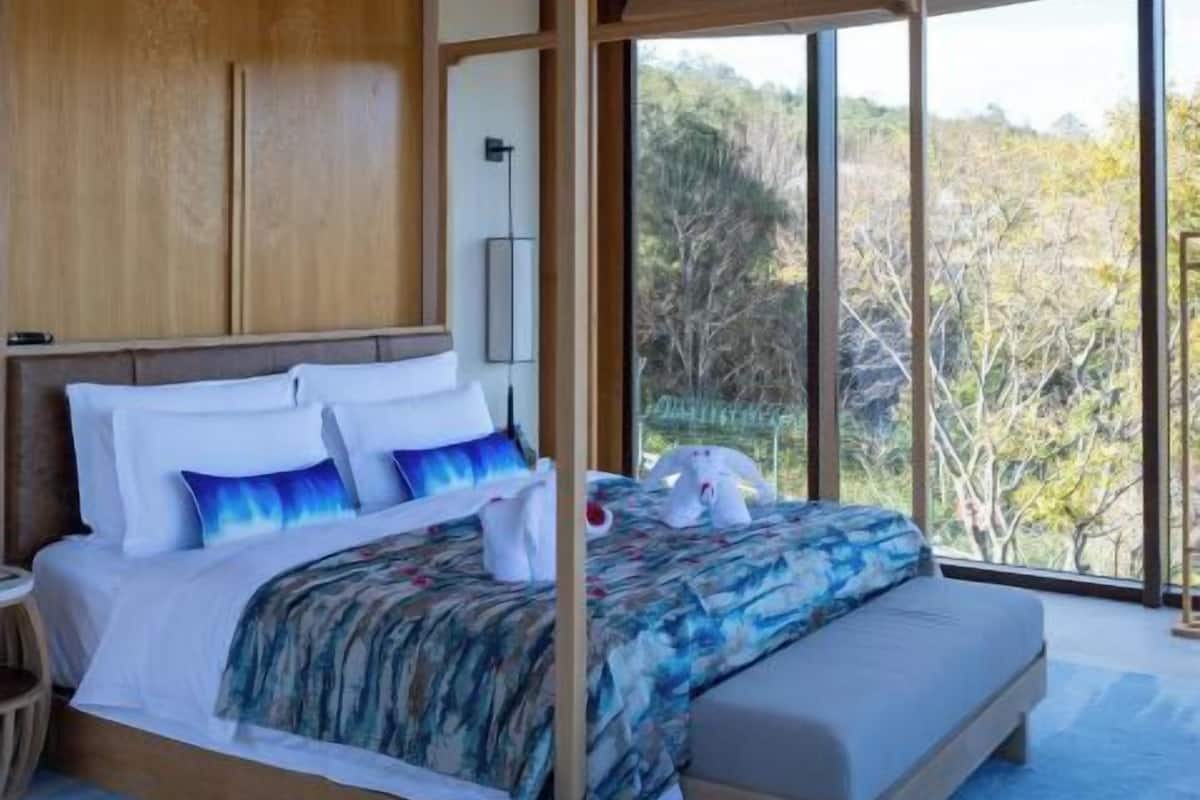 Un dormitorio moderno con una cama grande, cabecero de madera empotrado y vistas a los árboles a través de amplias ventanas.