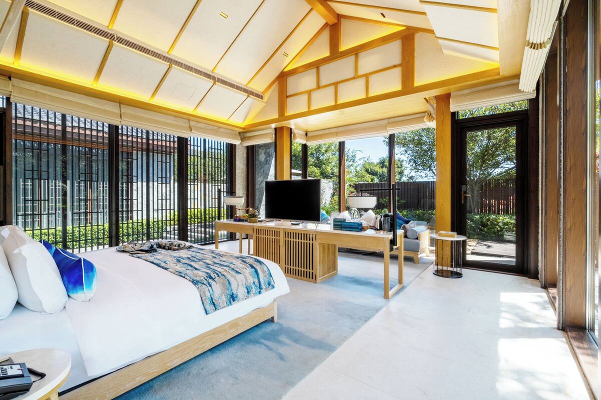 Un dormitorio moderno con cama, televisor, escritorio y ventanas amplias.