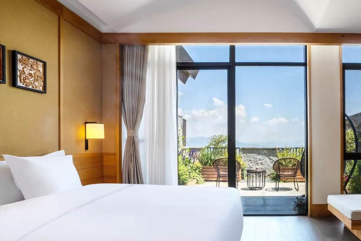 Habitación de hotel con una cama grande, una lámpara de noche, vistas al exterior y una ventana con cortinas.