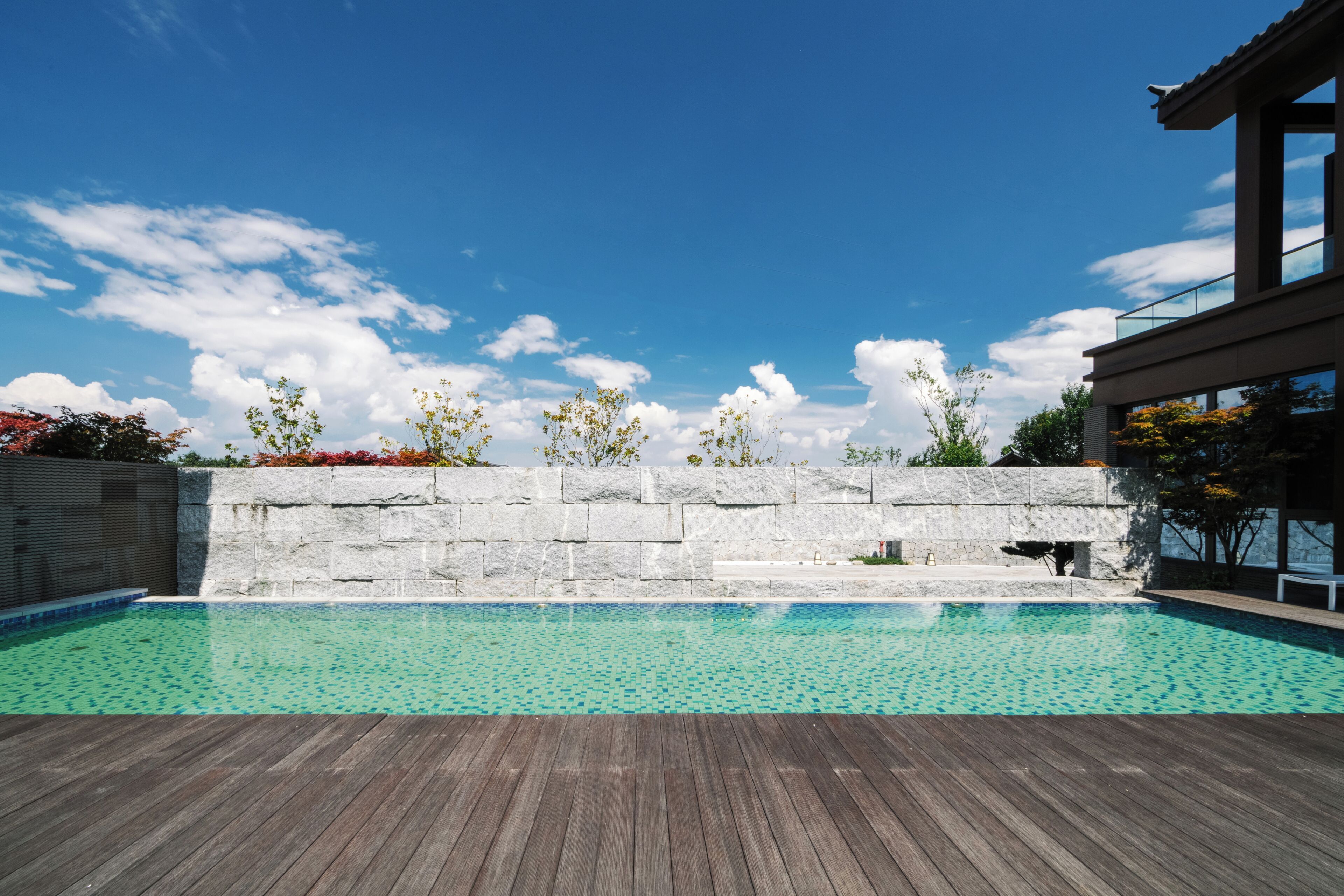 A swimming pool with a wooden deck, a stone wall, and a modern building in the background.