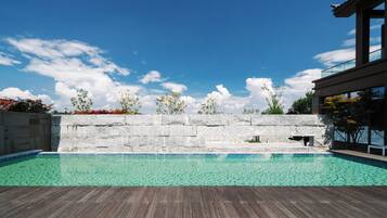 A swimming pool with a wooden deck, a stone wall, and a modern building in the background.