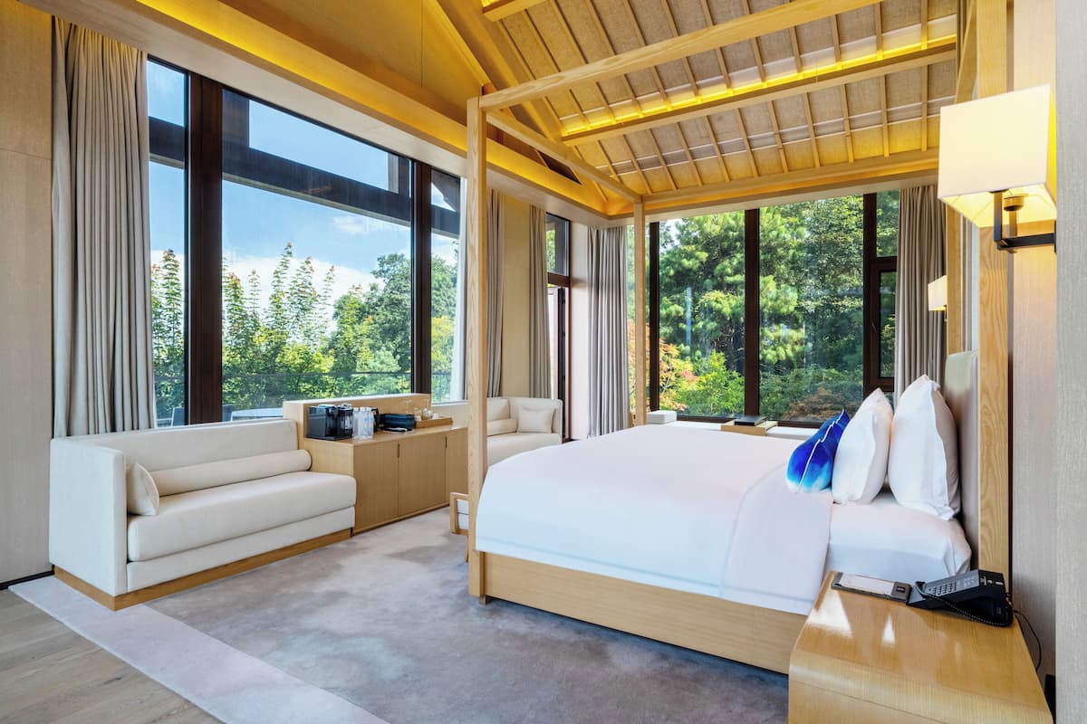 Una habitación de hotel moderna con una cama grande, un sofá, un escritorio con una cafetera y ventanas amplias que ofrecen vistas al exterior.