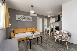 A modern living room with a sofa, a table with a remote control, a floral chair, a dining area with a table and chairs, and a kitchen with cabinets and a stove.
