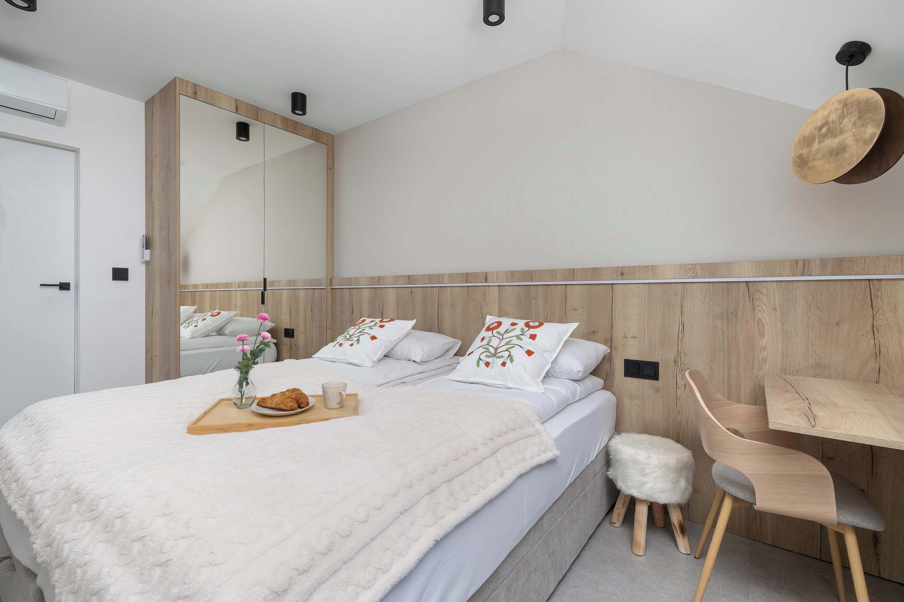 A modern bedroom with a large bed, bedside tables, a wooden headboard, a mirror, and a hanging light fixture.