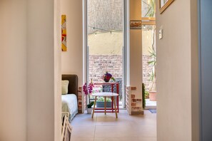 Ein schmales Flurzimmer mit einem Bett, einem kleinen Tisch mit Topfpflanze und Blick auf eine Backsteinmauer und einen Balkon.