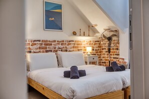 Ein Schlafzimmer mit Ziegelwand, einem Bett mit weißen Bettbezügen, einem Standventilator und einem Bild an der Wand.