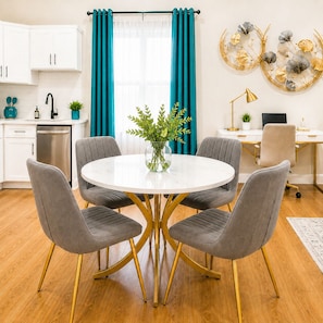 A modern dining area with a round table, four grey upholstered chairs, and a vase of green plants.