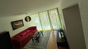 A living room with a red sofa, a coffee table, a painting on the wall, and a large window with sheer curtains. - A cozy room with double bed in semi shared Flat (Wallisellen)