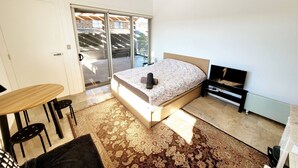 Ein Schlafzimmer mit einem Bett, einem kleinen Tisch, Stühlen, einem Fernseher und einer Schiebetür zum Balkon.