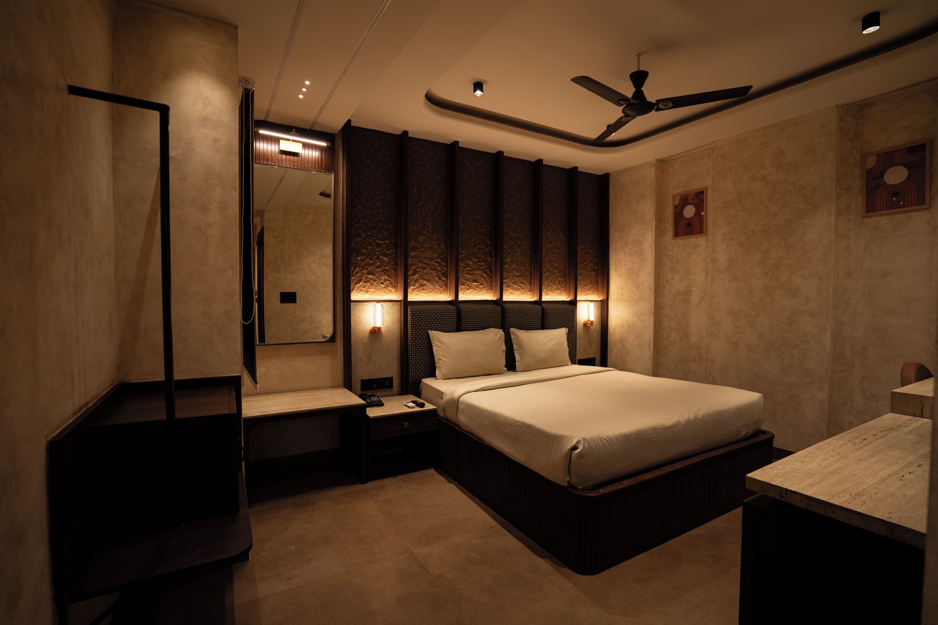 A modern hotel room with a bed, bedside tables, a nightstand, a ceiling fan, and wall-mounted lighting.
