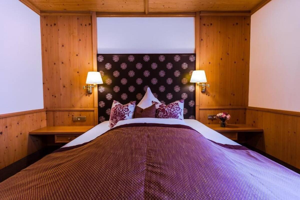 A hotel room with a wooden headboard, two bedside lamps, a bed with a patterned bedspread, and a nightstand with a vase of flowers.