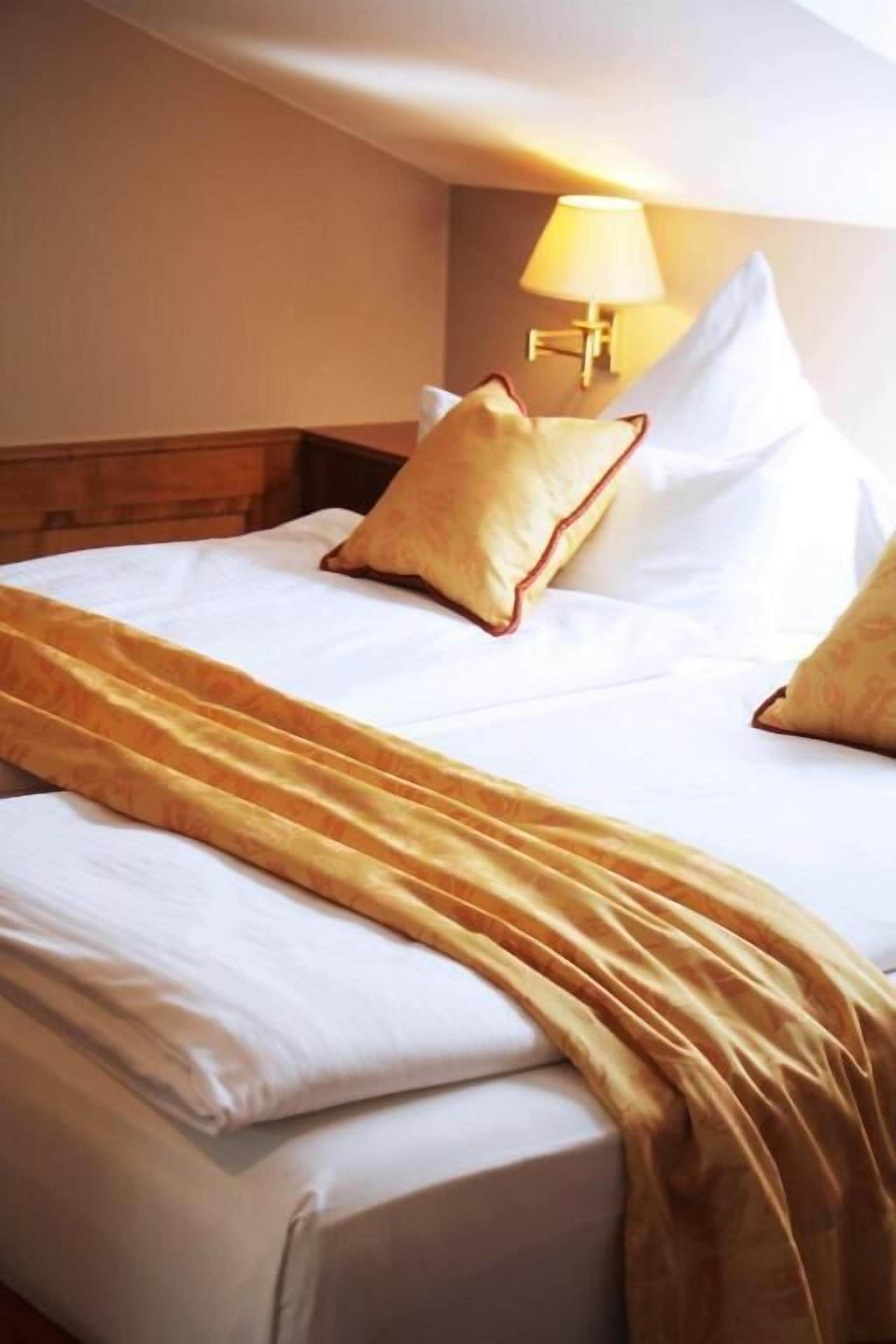 A neatly made bed with white linens and a golden throw blanket, a bedside lamp, and a wooden headboard.