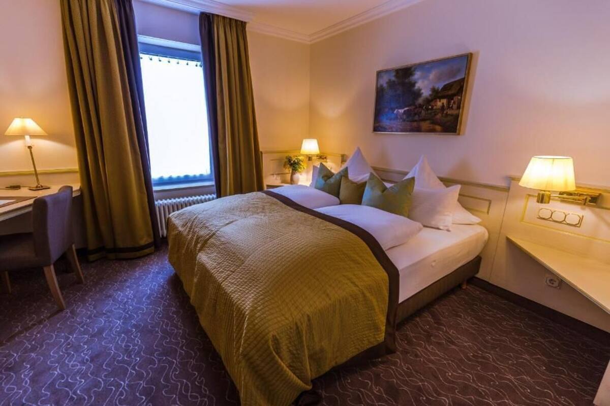 A hotel room with a bed, two bedside lamps, a desk, a chair, and a window with curtains.