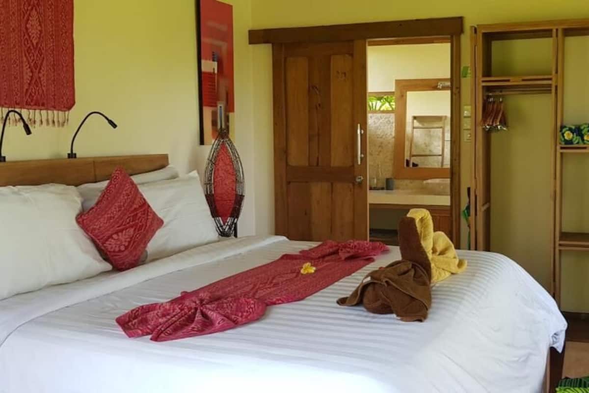 A bed with white linens, a red throw blanket, and a brown towel. There is a wooden door and a shelf with decorative items.