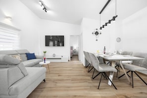 Ein modernes Wohnzimmer mit einem grauen Sofa, einem runden Couchtisch und einem Essbereich mit weißem Tisch und grauen Stühlen.
