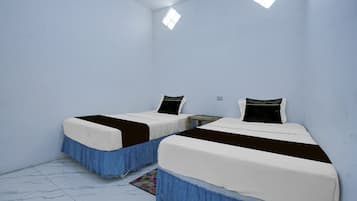 Habitación con dos camas, cada una con un colchón blanco y un cabecero oscuro. Hay una pequeña mesa entre las camas y las paredes son azul claro, con suelo de baldosas.