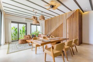 A modern dining area with a long wooden table, beige chairs, pendant lighting, and a slatted wooden partition. - Siddhi Villa (Kerobokan)