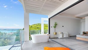 A modern bathroom with a freestanding bathtub, a glass-enclosed balcony, and a view of a coastal landscape. - Luxury Sunset House Home Gym Cinema (Bophut)