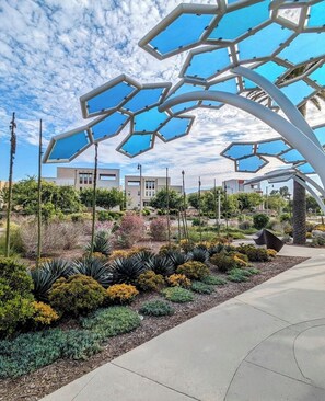 Un jardín ajardinado con una escultura metálica de diseño geométrico, un camino empedrado y edificios modernos al fondo.