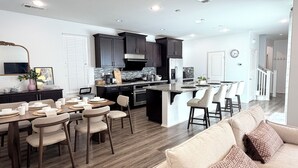 A modern kitchen with dark cabinets, a central island, and a dining area with chairs and a table. - Irvine Great Park Beacon 3 B &4 B two story house (Irvine)