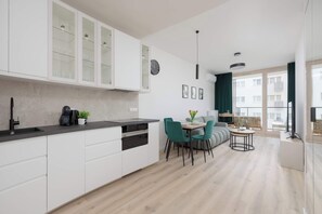Eine moderne Küche mit weißen Schränken, eine Essecke mit türkisfarbenen Stühlen und Blick auf ein Gebäude durch große Fenster.