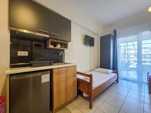 A compact kitchen with a refrigerator, microwave, and sink. A bed is positioned near a large window. - 104 - EVROSTAR Studio by SDR (Paralia)