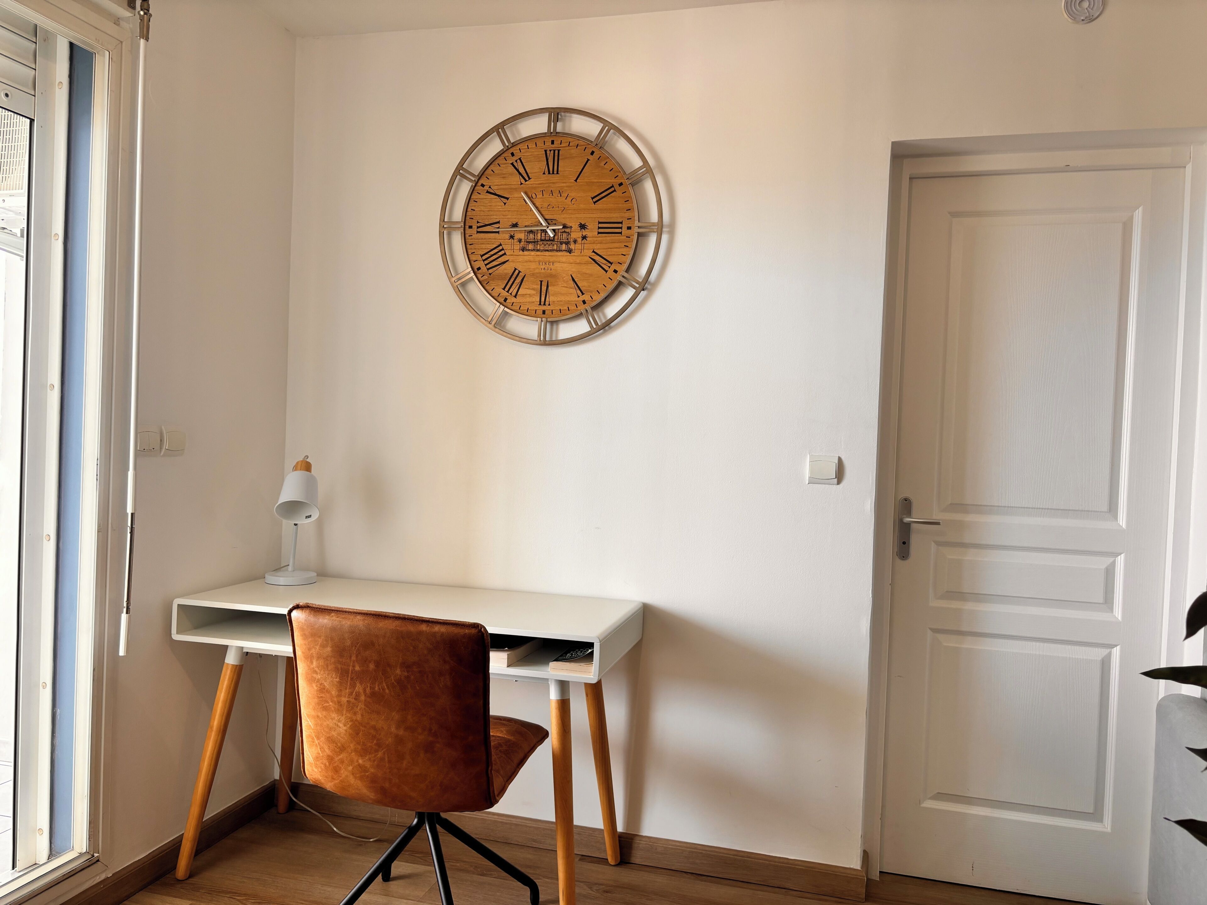 Um quarto com um relógio de parede de madeira, uma escrivaninha branca, uma poltrona de couro marrom e uma porta branca.