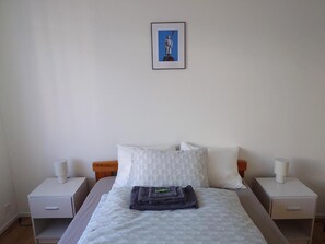 Ein Hotelzimmer mit einem Bett, zwei Nachttischen, einem kleinen Tisch, einem gerahmten Bild an der Wand und einem auf dem Bett gefalteten Handtuch.