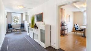 Una sala moderna con un sofá blanco, una mesa de centro y un televisor sobre un mueble blanco. Hay una zona de comedor con una mesa y sillas, y una estantería con objetos decorativos.