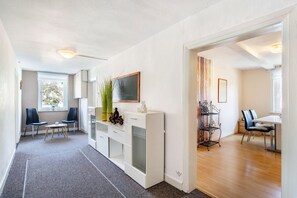 Un salon moderne avec un canapé blanc, une table basse et un téléviseur posé sur un meuble blanc. On y trouve également un coin repas avec une table et des chaises, ainsi qu’une bibliothèque agrémentée d’objets décoratifs.