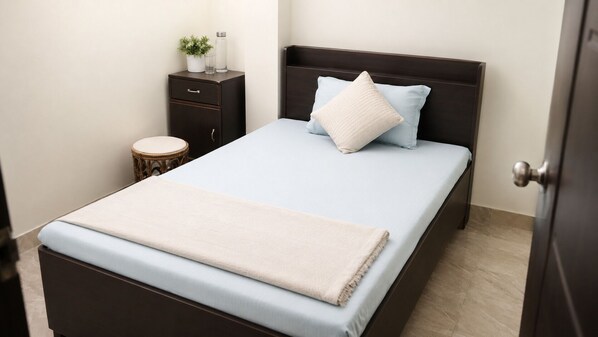 Una cama bien tendida con colchón azul claro y manta color beige, cabecera de madera oscura, mesita de noche con una planta y un vaso, y un taburete redondo de madera.