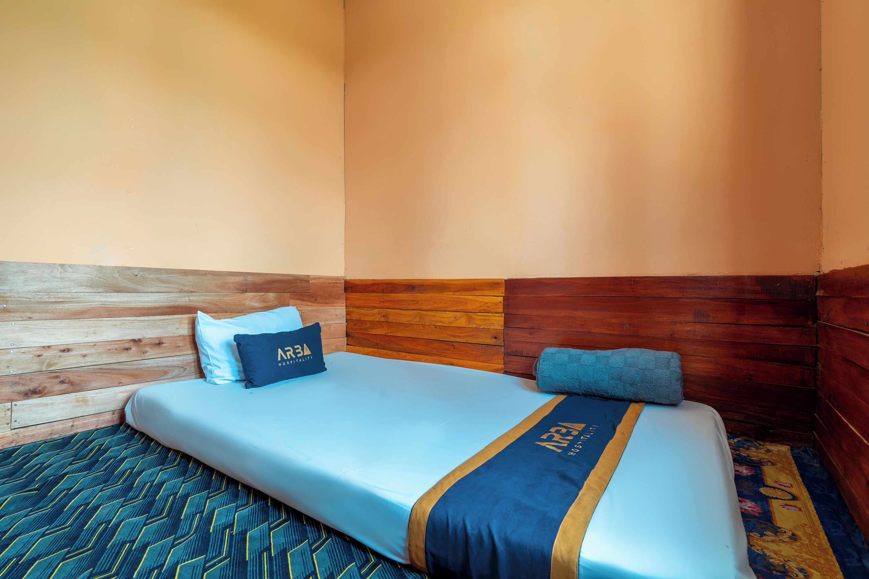 A single bed with a blue and gold bedspread, a pillow with 'ARA' text, and a wooden headboard.