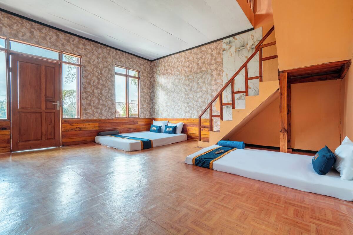 A spacious room with a wooden floor, a large window, a staircase, and a bed with pillows.