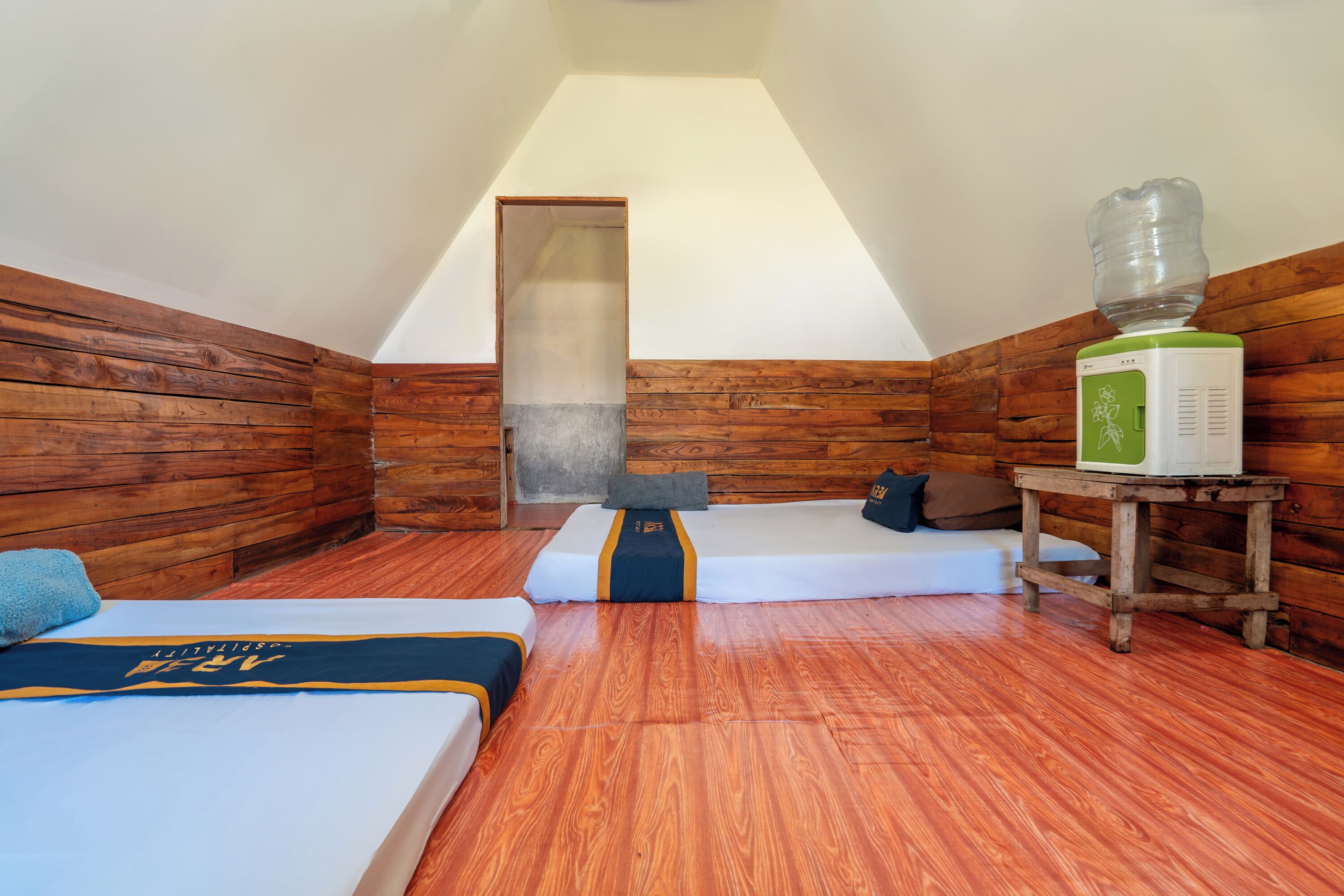 A room with wooden walls, a sloped ceiling, a bed with a blue and yellow bedspread, a small table with a water dispenser, and a bench with pillows.