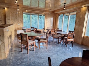 Una habitación con paneles de madera, mesas y sillas redondas, ventanas amplias y suelo de piedra.