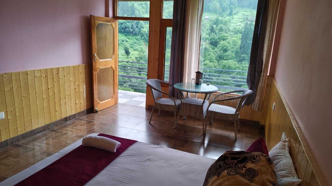 Quarto com cama, mesa e cadeiras, vista para o verde lá fora e porta de madeira.