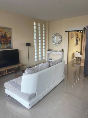 A modern living room with a white sofa, a flat-screen TV, a large window with vertical blinds, and a decorative mirror. - Romantic Tropical Villa Retreat in Maret, Thailand (Maret)