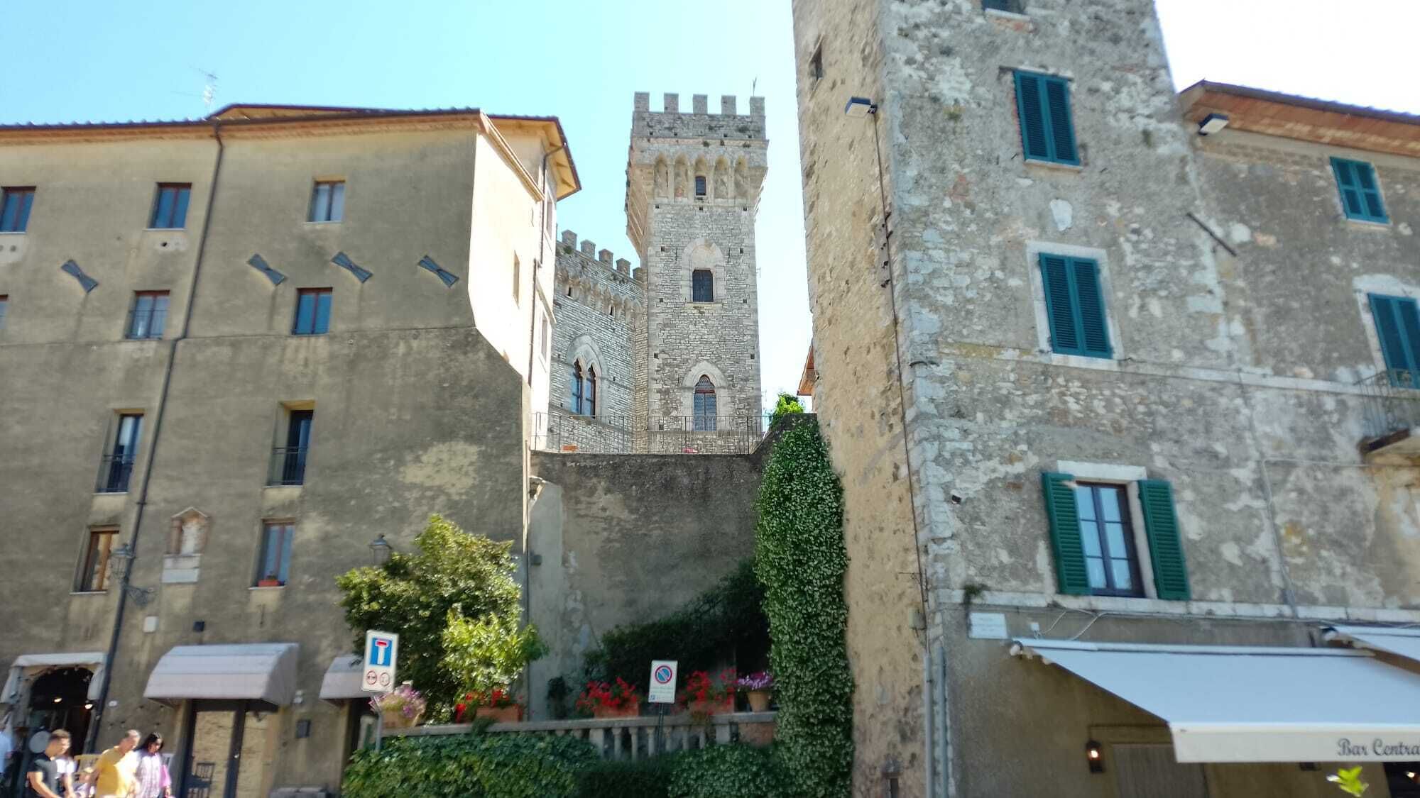 Une tour de pierre historique, surmontée de créneaux, des bâtiments adjacents dotés de balcons et un petit bar avec des places assises en plein air.