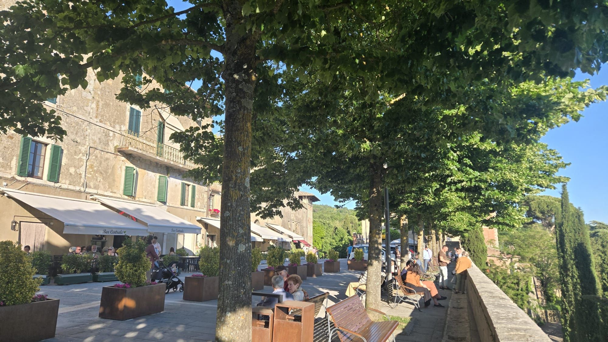 A tree-lined street with outdoor seating, buildings with shutters, and a clear sky.