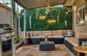 Eine Terrasse im Freien mit Grill, einer Sofagruppe und einer Wand mit Pflanzen und einem Neonlicht.
