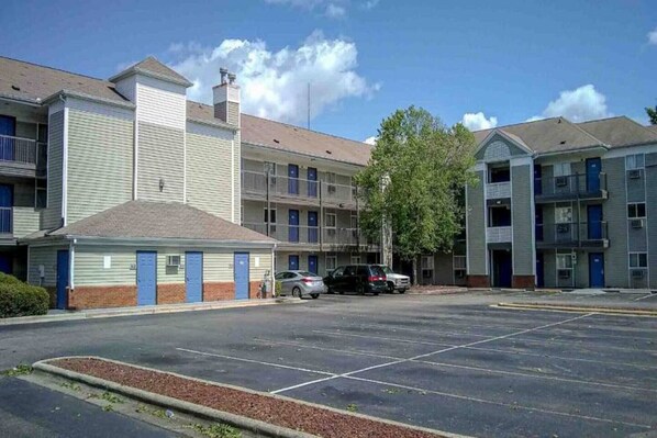 Un motel à plusieurs étages avec un parking, un petit bâtiment doté d’un toit en pente et un arbre devant.